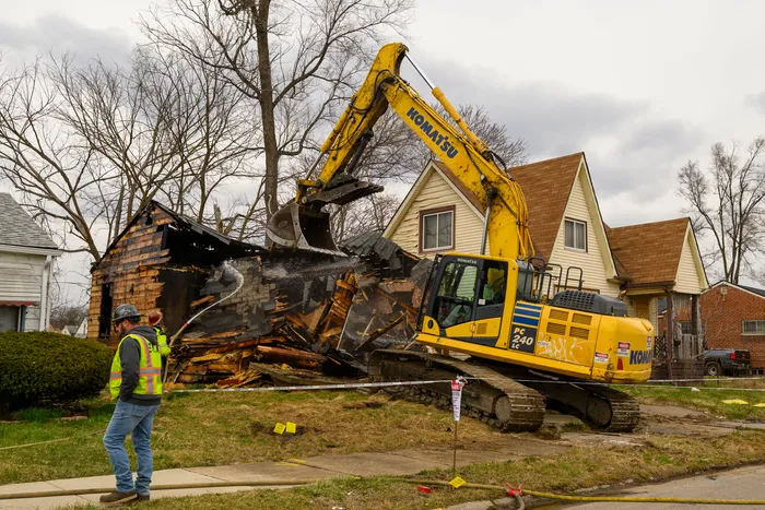 Yellow Komatsu excavator tearing down a burned house while a worker in a safety vest looks on