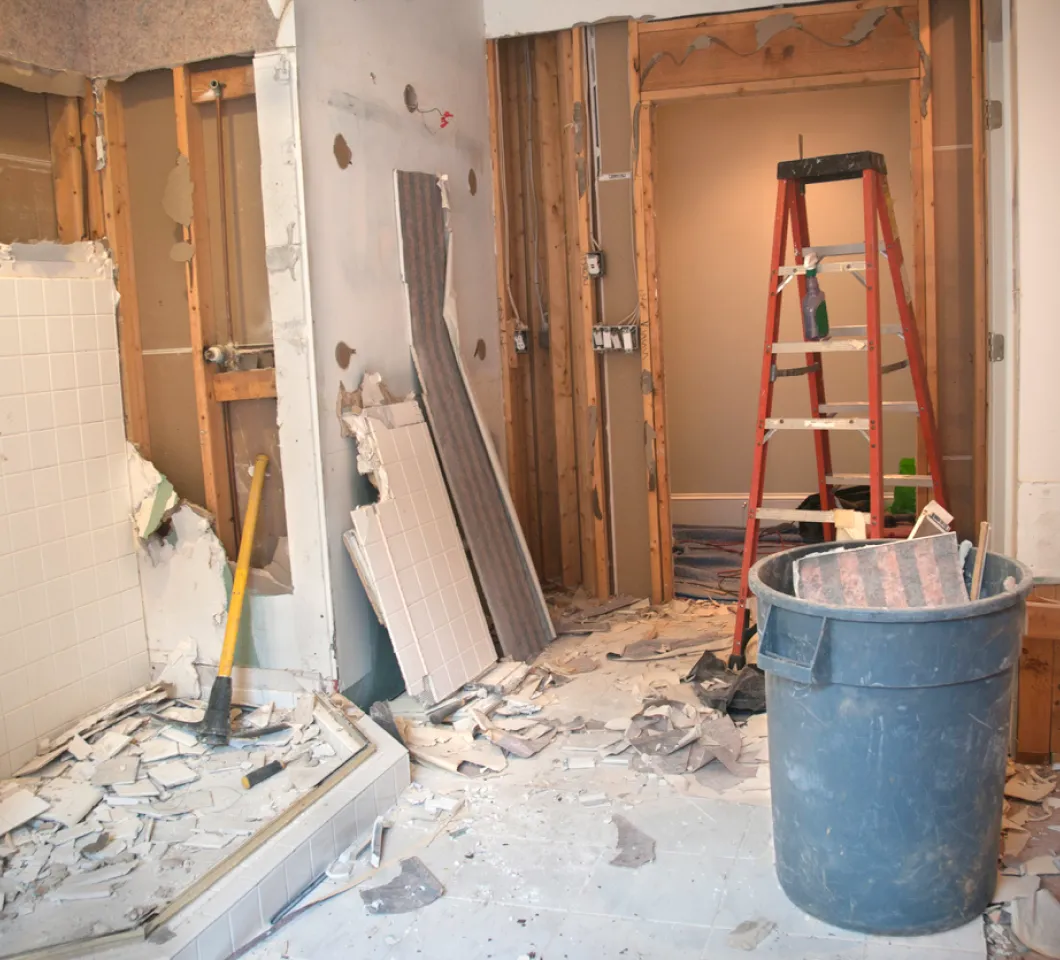 Gutted bathroom with broken tile, exposed wall studs, a ladder and a trash can